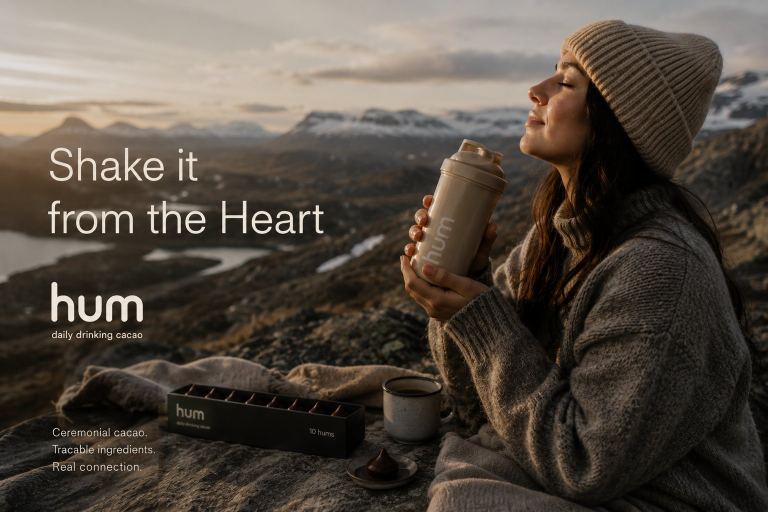 A woman in a knit beanie holding a warm HUM shaker on a mountainside at golden hour